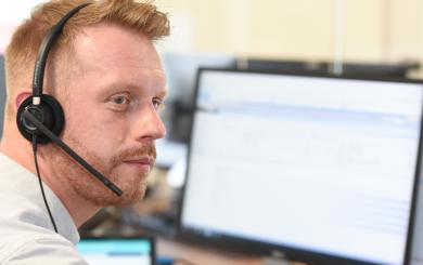 A ginger haired man with a beard wearing a phone headset. There are computer screens on a desk visible in the background.