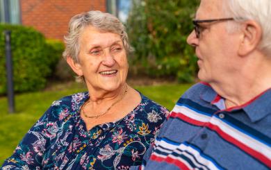 Two elderly individuals are sitting outdoors on a bench, facing each other. One wears a dark floral blouse with a gold necklace, and the other wears a blue polo shirt with red, white, and blue stripes. They appear to be engaged in conversation. The background includes greenery and part of a brick building. 