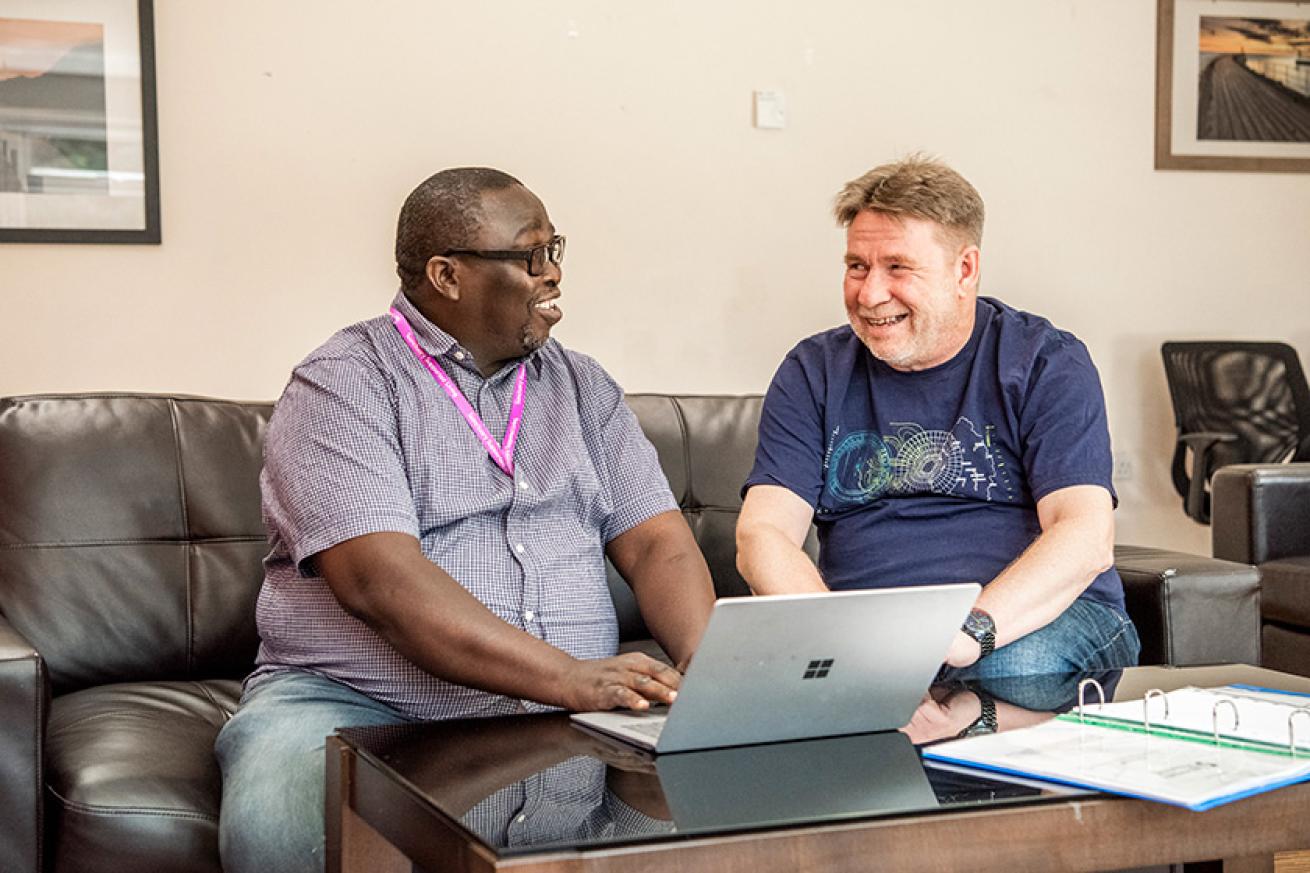 Supported service user and member of staff at Stepping Stone Court A supported service user and a member of staff sat together and smiling, they are sat on a brown leather sofa, in front of a laptop on top of a coffee table.
