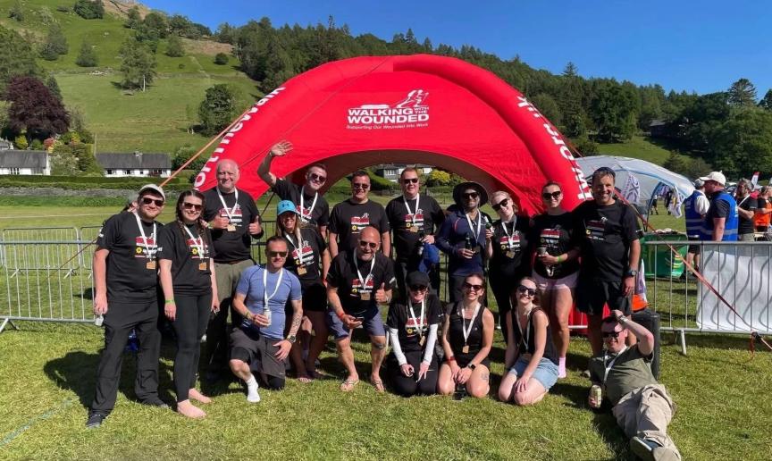 A group of people wearing black 'Walking With The Wounded' t-shirts pose in front of a red inflatable arch with the same logo. They are outdoors on grass with hills and trees in the background. Some are standing, kneeling, or sitting, and many are holding medals.