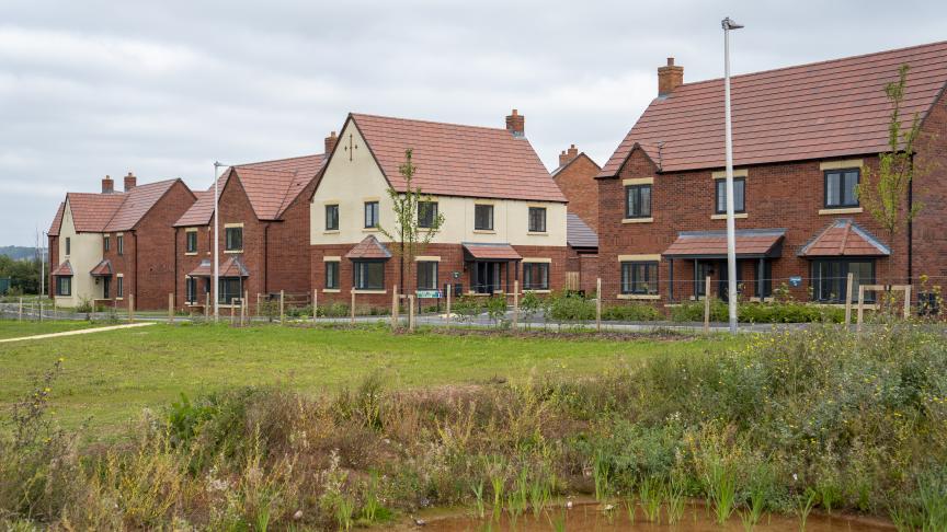 A Sanctuary new-build development estate, build with red brick and cream rendering. There is a large grassy area in the forefront of the image