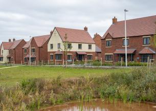 A Sanctuary new-build development estate, build with red brick and cream rendering. There is a large grassy area in the forefront of the image